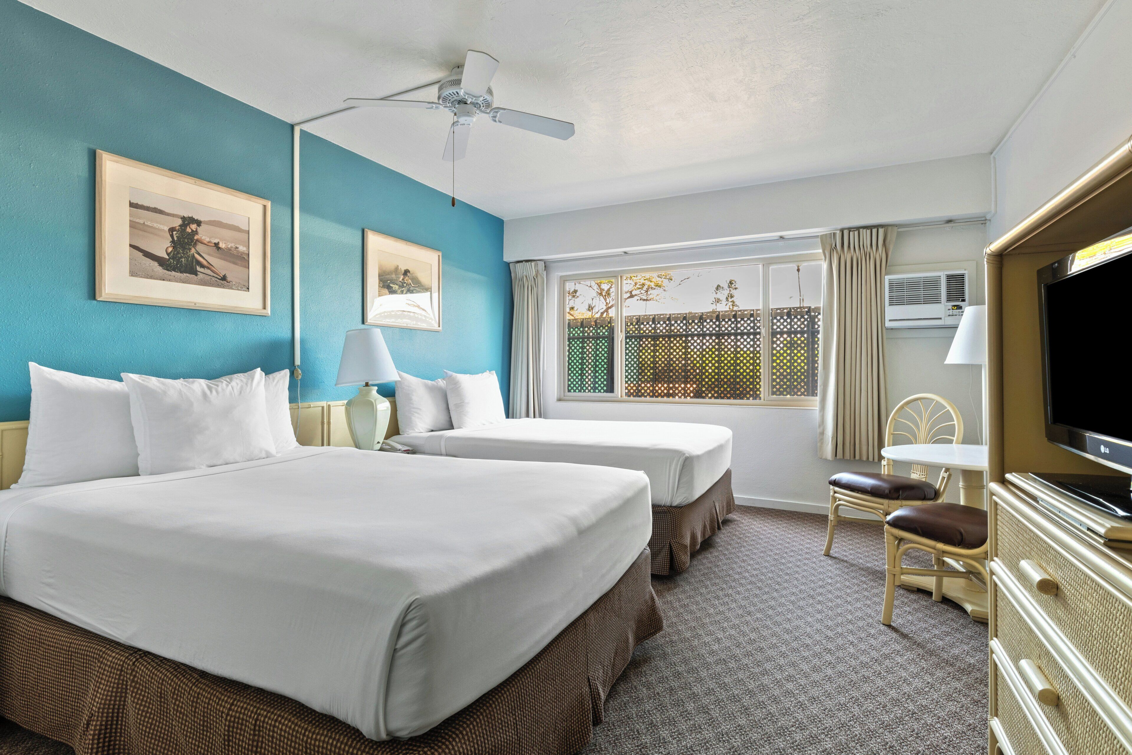 A hotel room with two beds, a ceiling fan, a TV, a window with a view of greenery, and a dining area with a table and chairs.