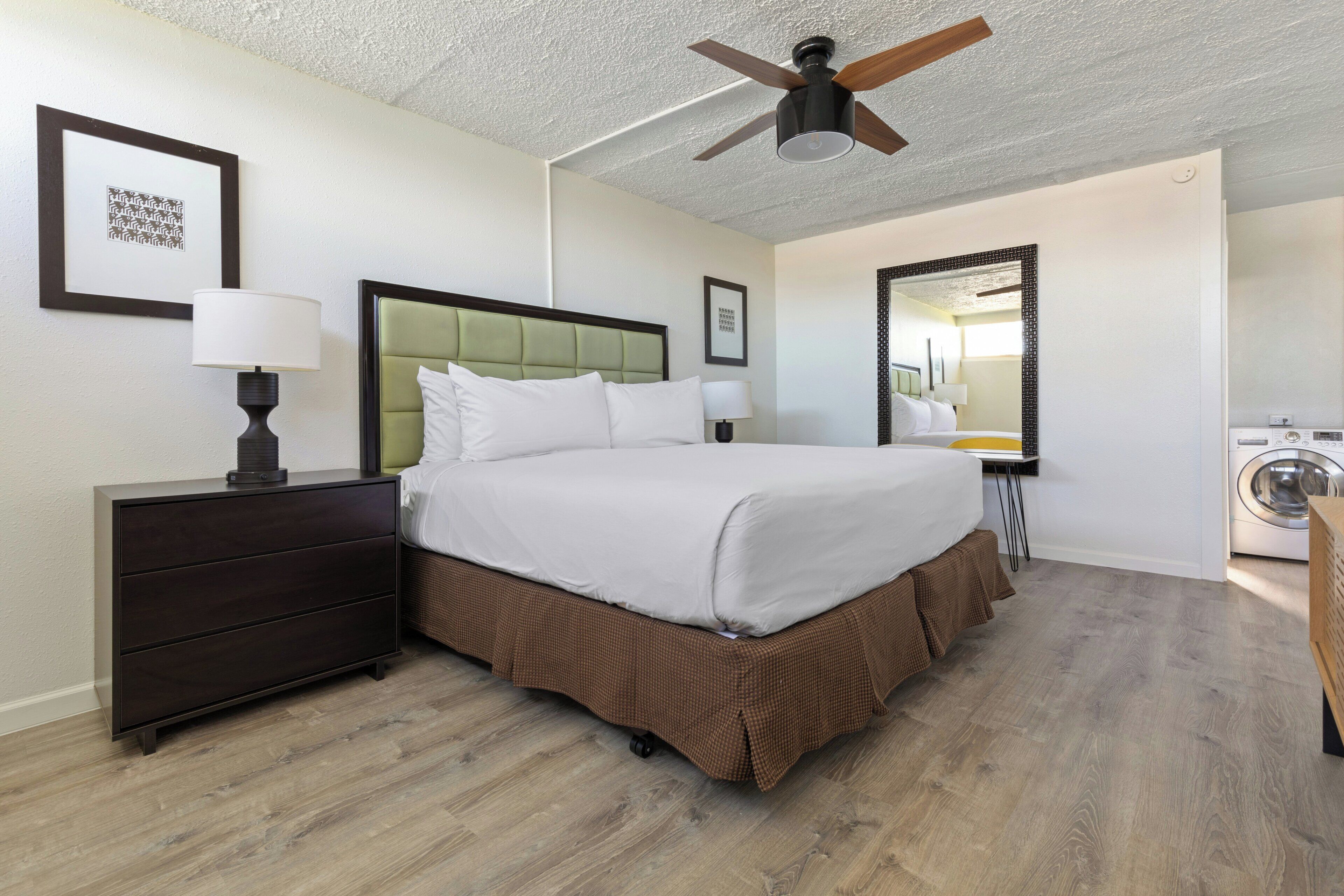 Habitación de hotel con cama, mesitas de noche, ventilador de techo, espejo y lavadora.