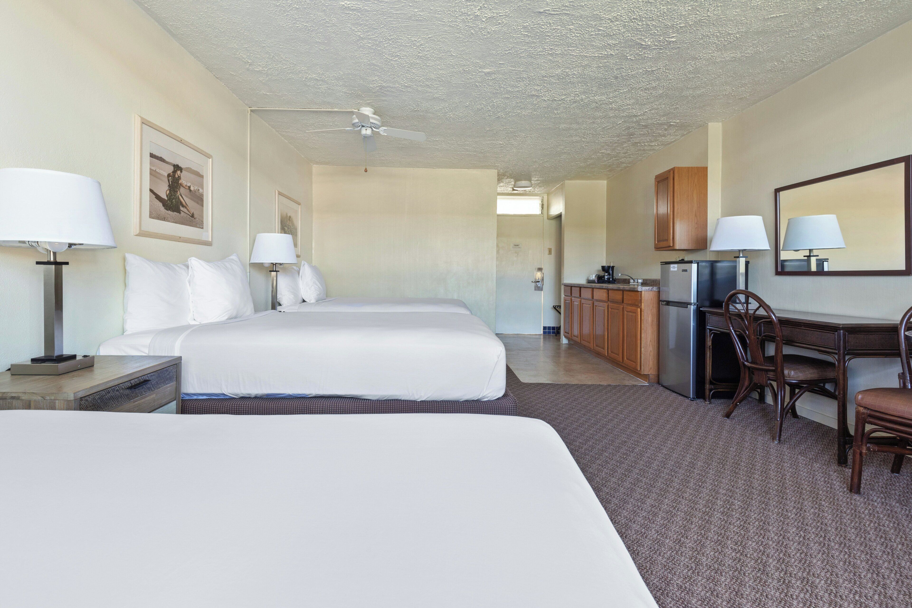 Habitación de hotel con cama, escritorio, silla, refrigerador y ventilador de techo.