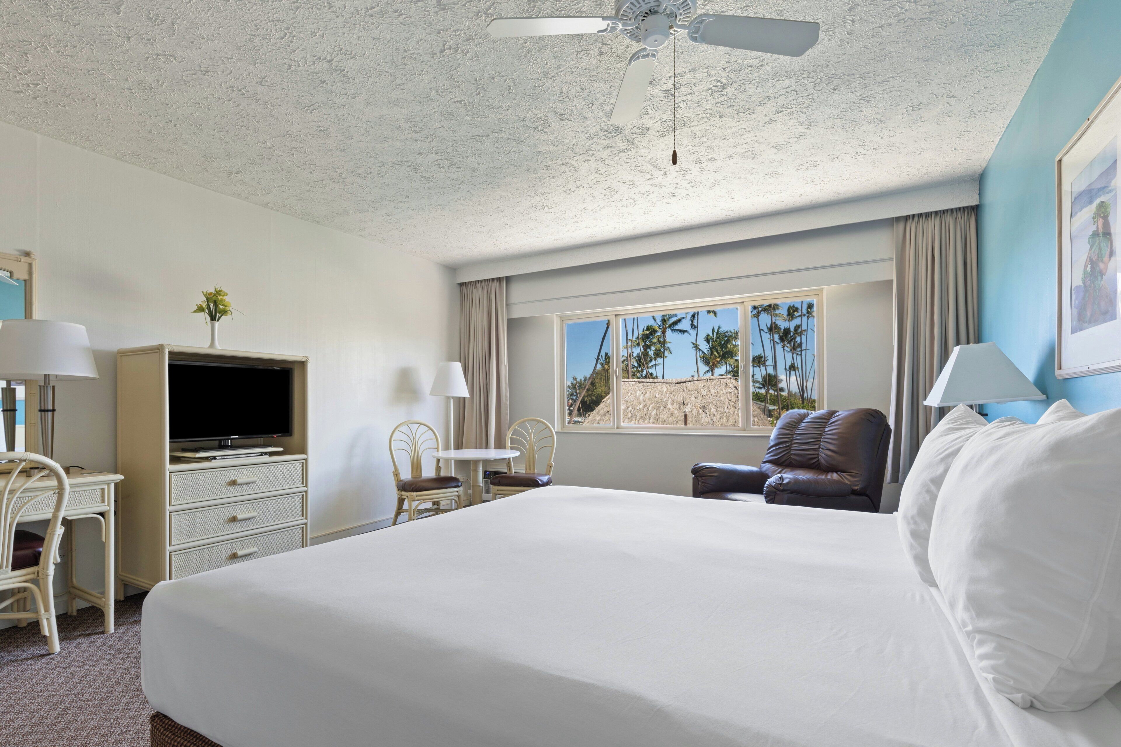 A hotel room with a bed, a desk, a television, a chair, a window with a view, and a ceiling fan.