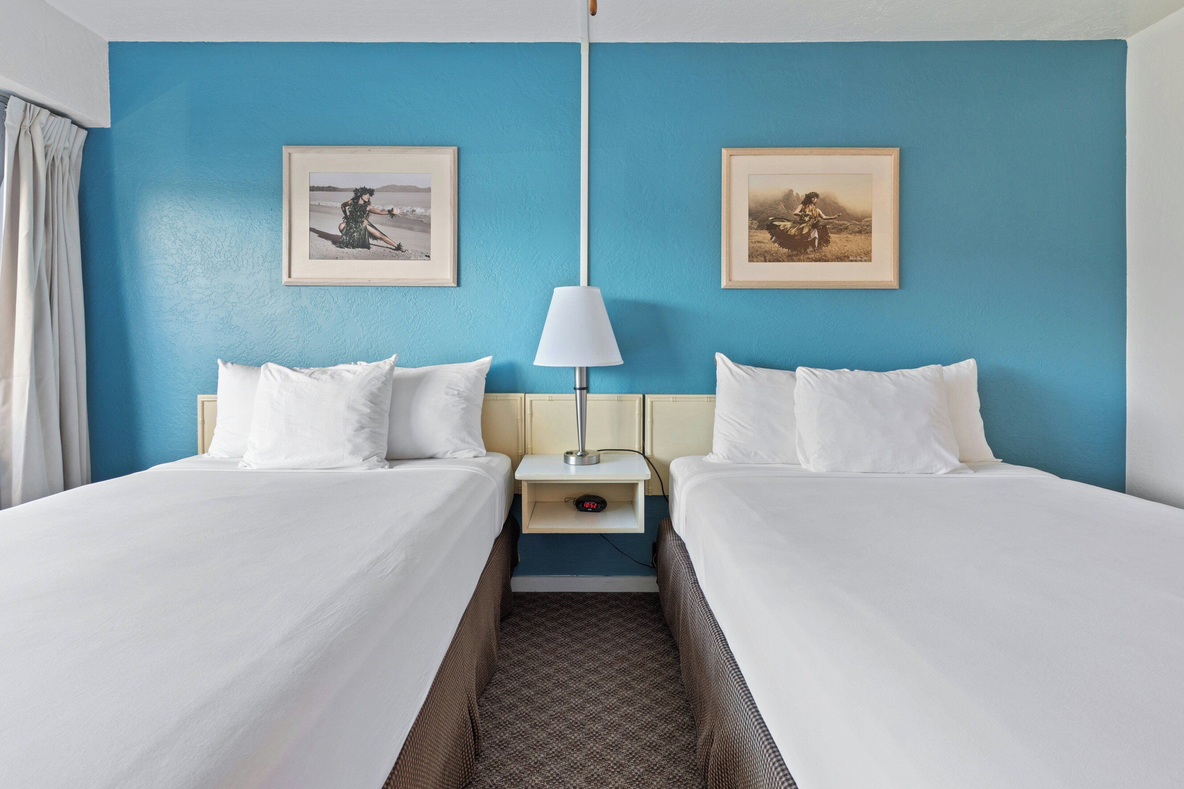 Habitación de hotel con dos camas, una mesita de noche con lámpara y dos cuadros en la pared.