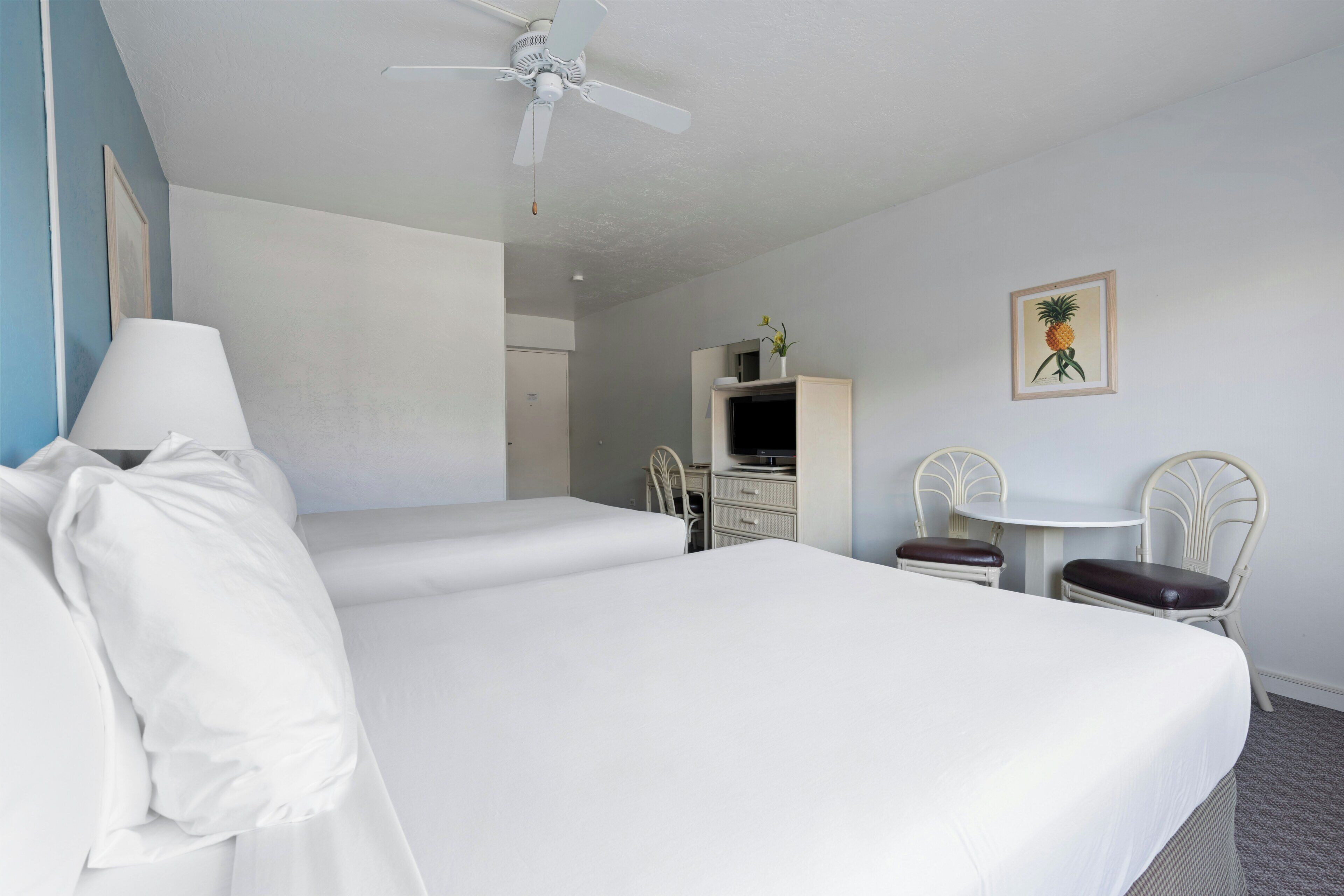 A hotel room with a bed, a desk, a chair, a small table, and a ceiling fan.