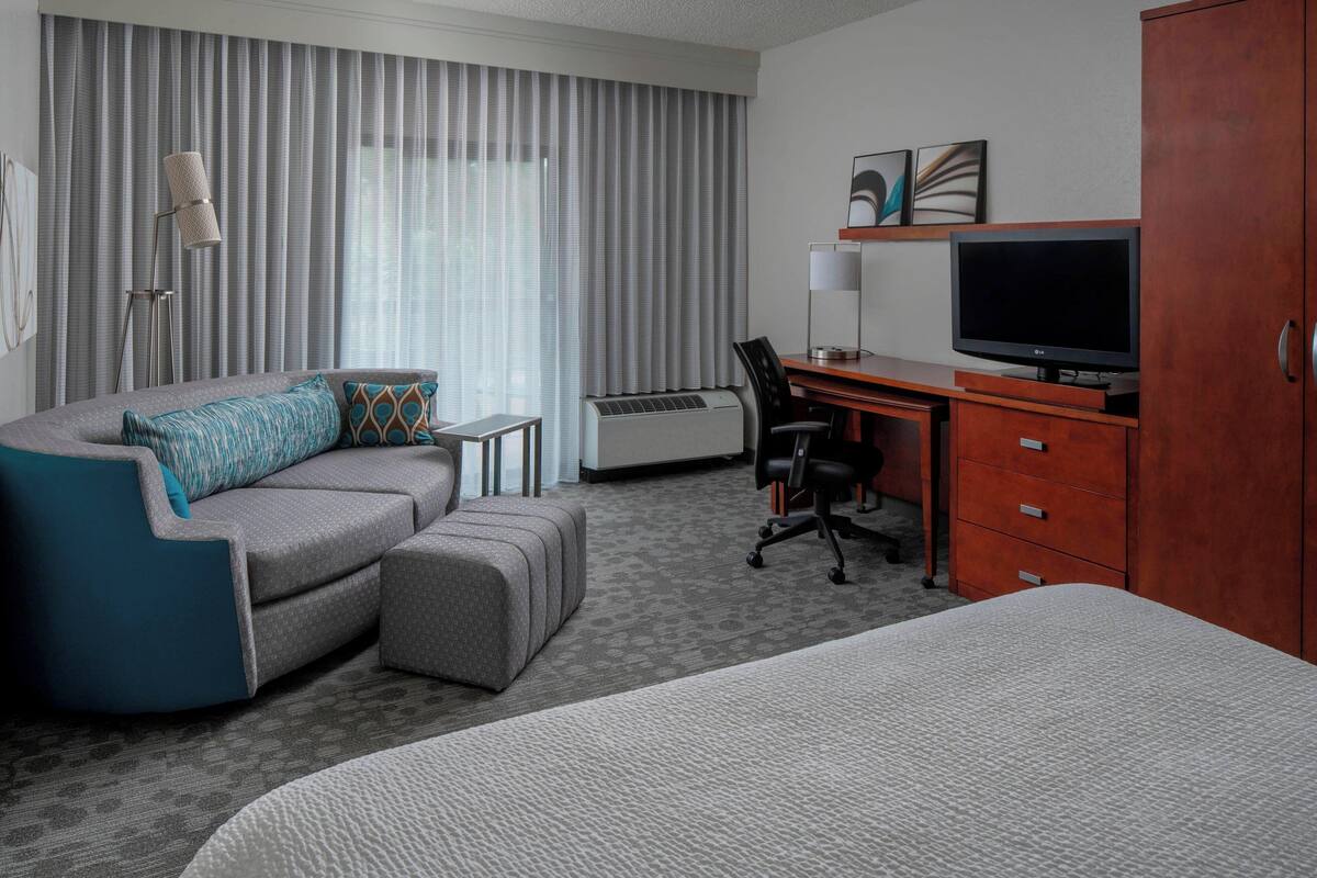 Une chambre d’hôtel avec un canapé gris, un petit bureau avec une télévision, une chaise et un grand lit.