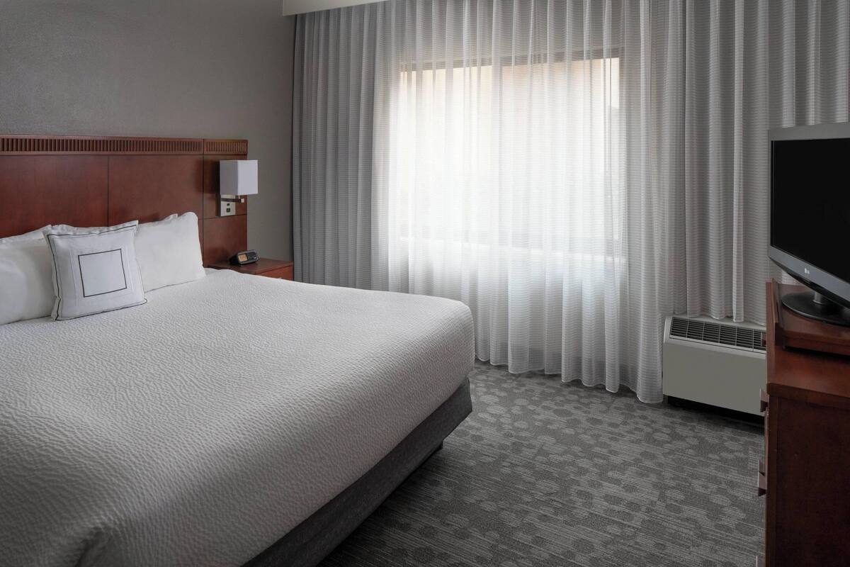 A hotel room with a large bed, a flat-screen TV, a bedside table, and a window with sheer curtains.
