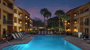 A hotel pool area with lounge chairs and umbrellas, surrounded by multi-story buildings with balconies. - Courtyard by Marriott Jacksonville at Mayo Clinic Campus/Beaches (Jacksonville)