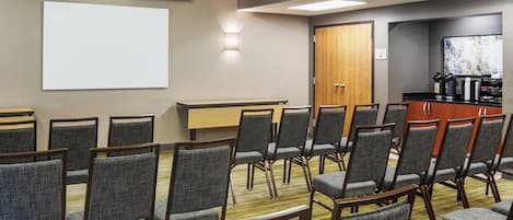 A conference room with rows of chairs, a projector screen, a coffee station, and a wooden door.