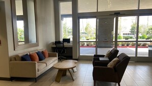 Eine großzügige Lobby mit einer Couch, Sesseln, einem kleinen Tisch und einem Schreibtisch mit einem Computer. Große Fenster bieten einen Blick ins Freie.
