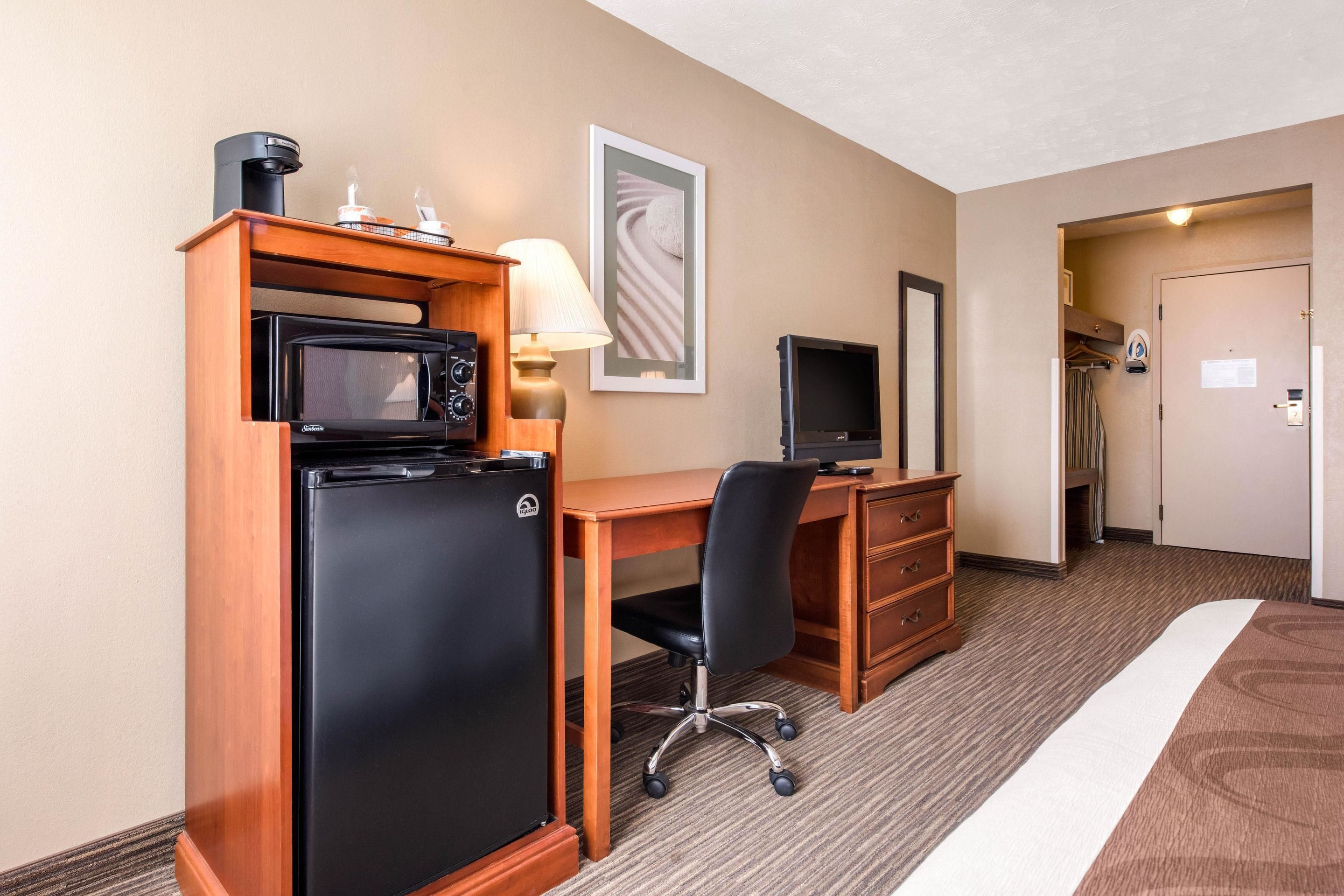 A hotel room with a wooden desk, a black refrigerator, a television, and a bed with a brown headboard.