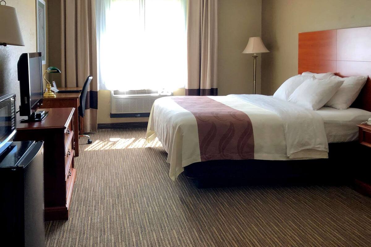 A hotel room with a bed, a desk, a television, and an air conditioning unit.