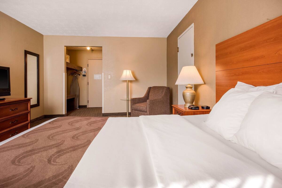 A hotel room with a large bed, a desk with a lamp, a chair, a TV, and a wardrobe.