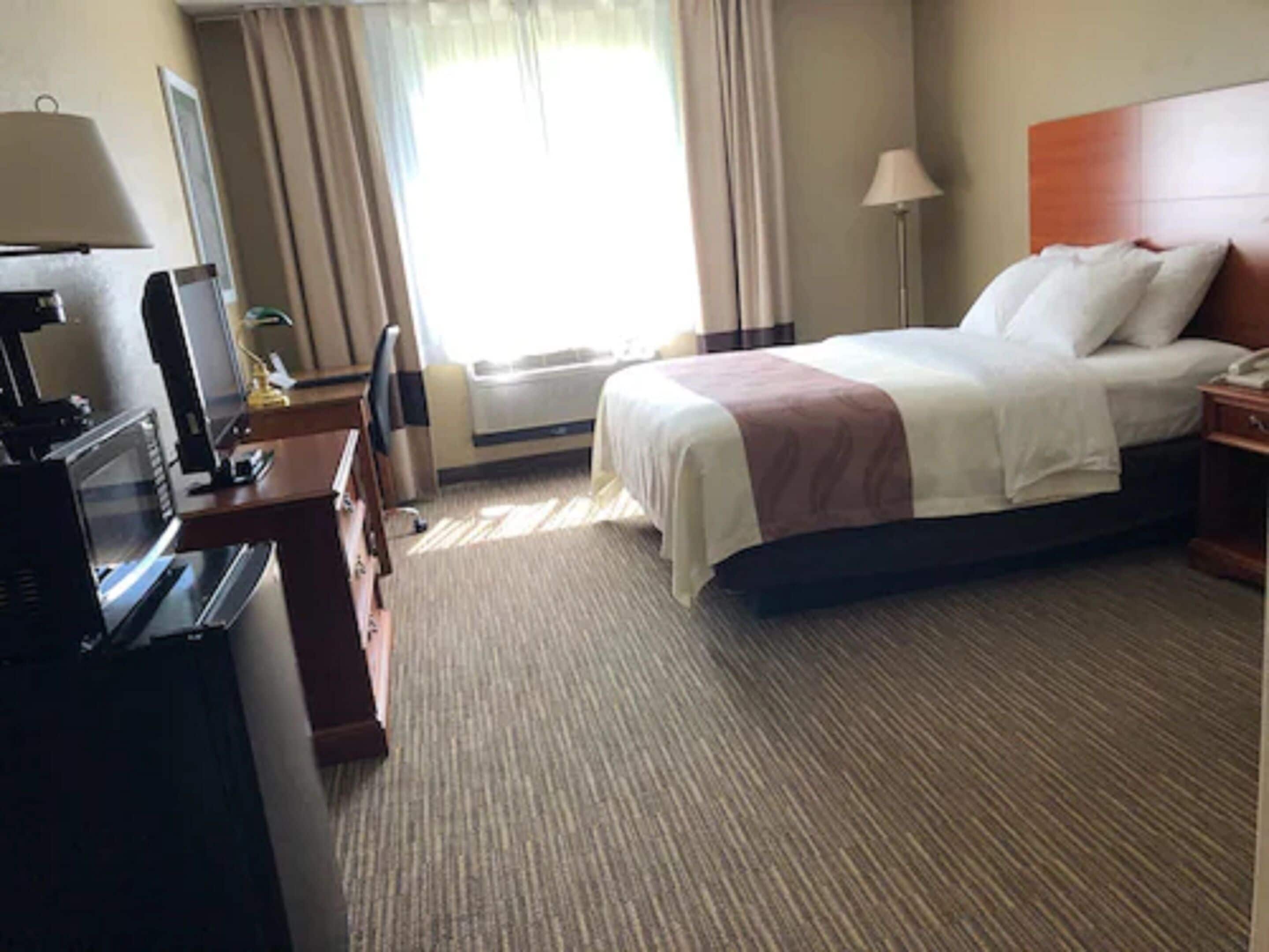 Une chambre d’hôtel avec un lit, un bureau avec une lampe, une télévision et une fenêtre avec des rideaux.