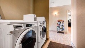 A laundry room with two front-loading washing machines and a wall-mounted dryer. There is a bulletin board with various items pinned to it.