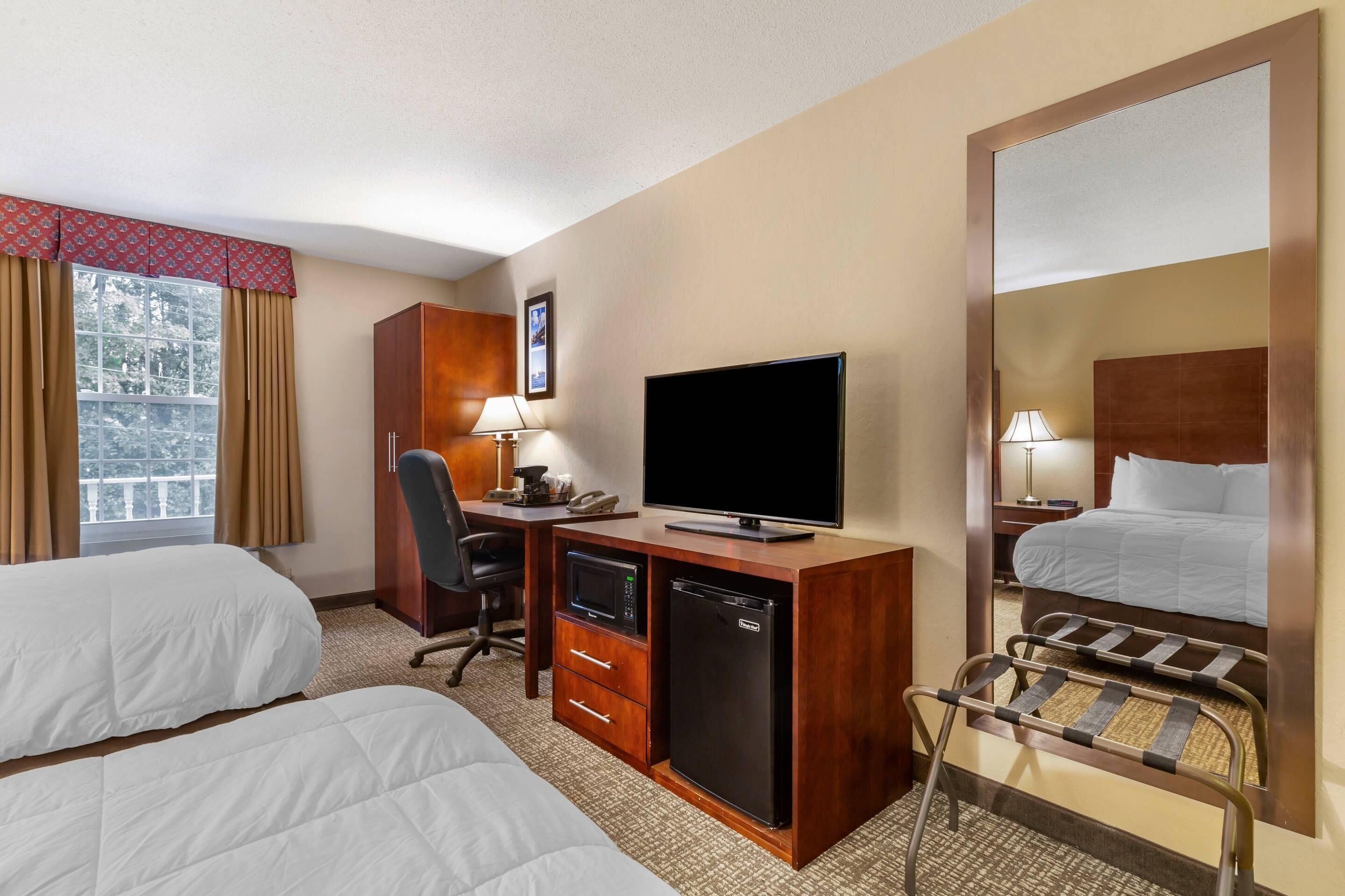 A hotel room with two beds, a desk with a TV, a microwave, a mini-fridge, a chair, and a large mirror.