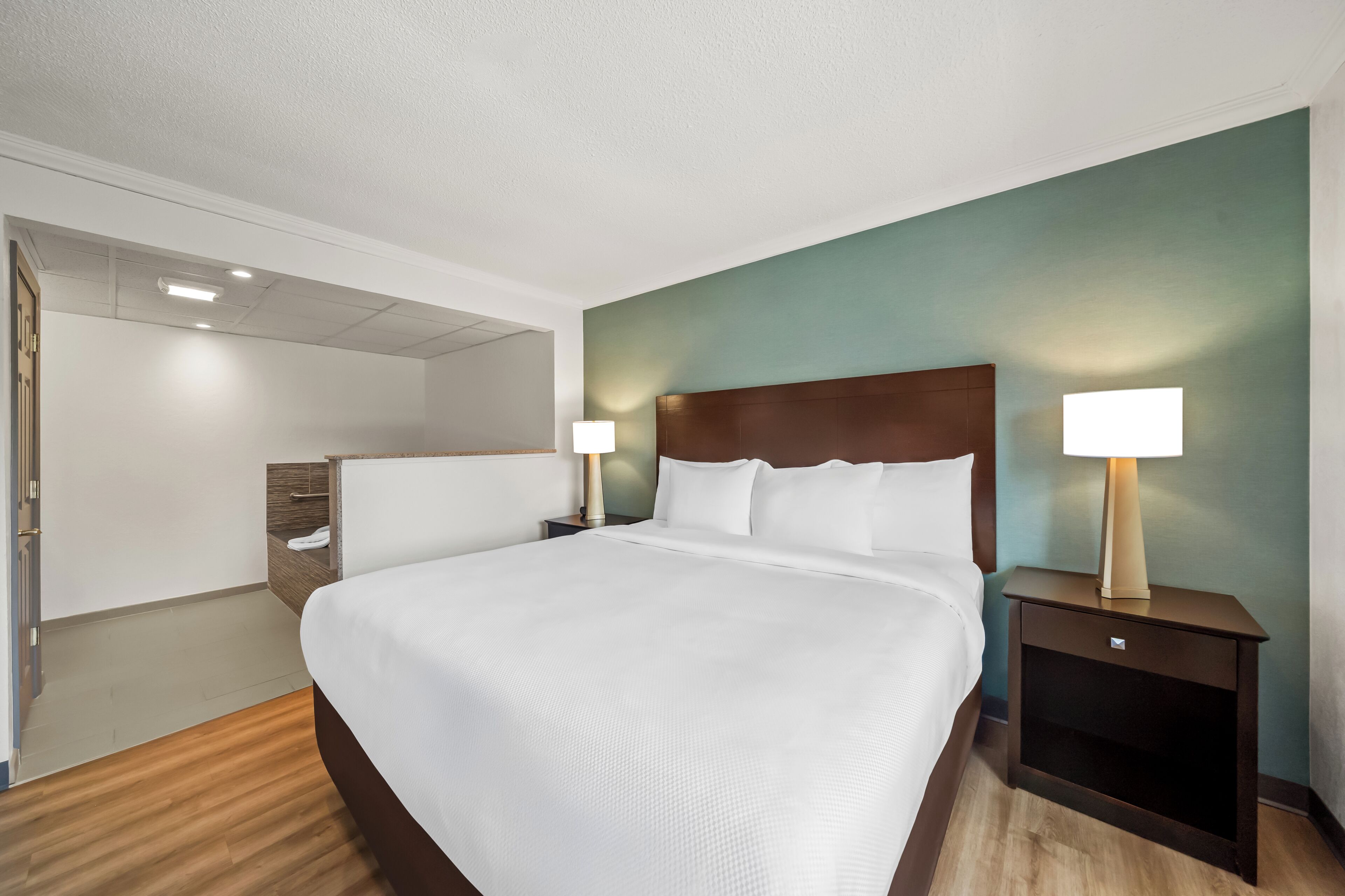 A hotel room with a large bed, two bedside tables with lamps, a wooden nightstand, and a wall-mounted shelf.