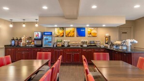 A hotel dining area with a long wooden table, red upholstered chairs, a coffee station, and wall art depicting food.