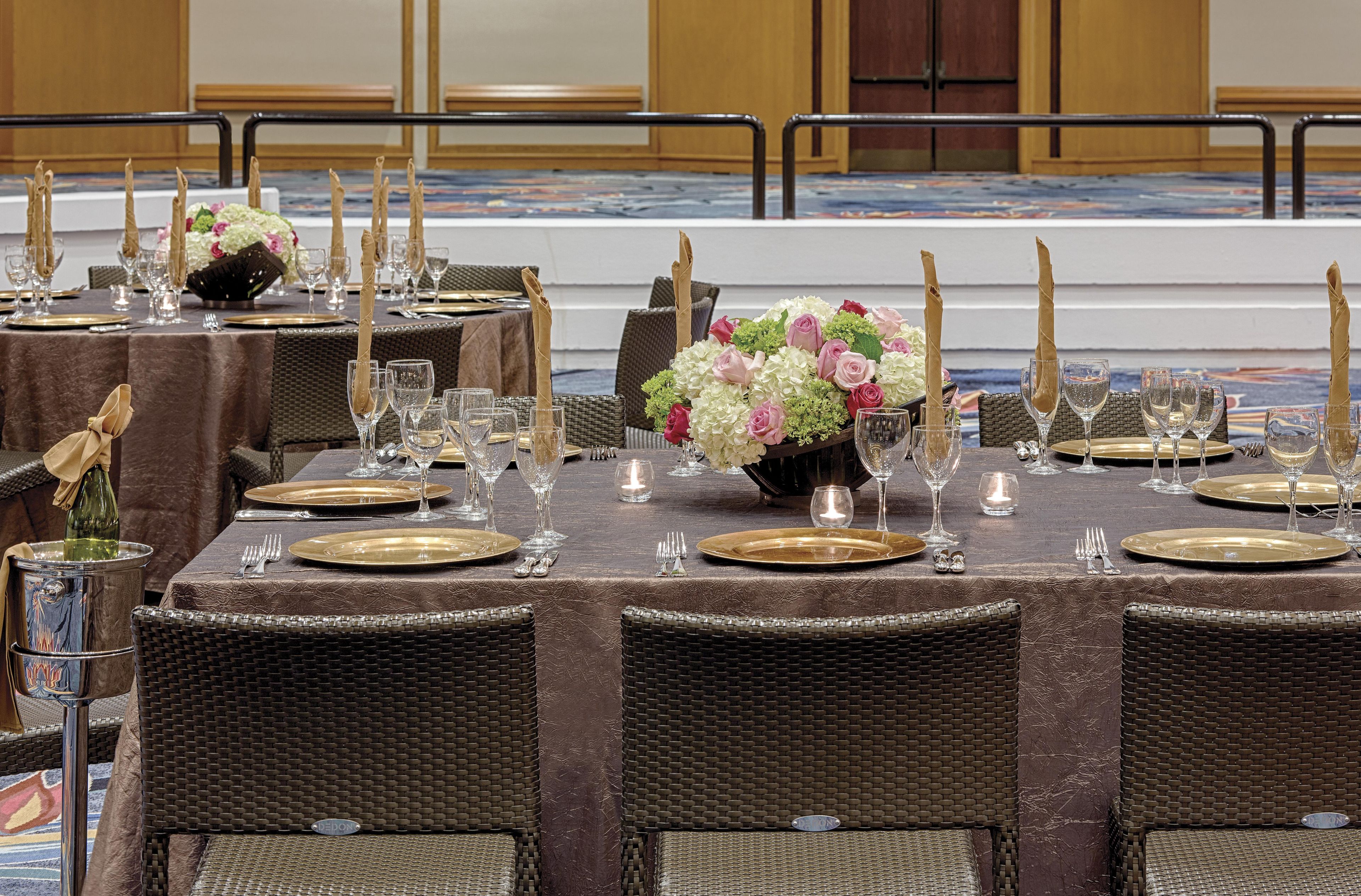 A banquet hall with tables set for a formal event, featuring wicker chairs, gold-rimmed plates, and floral centerpieces.