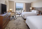A hotel room with two beds, a desk with a television, a chair, and a view of the outdoors.