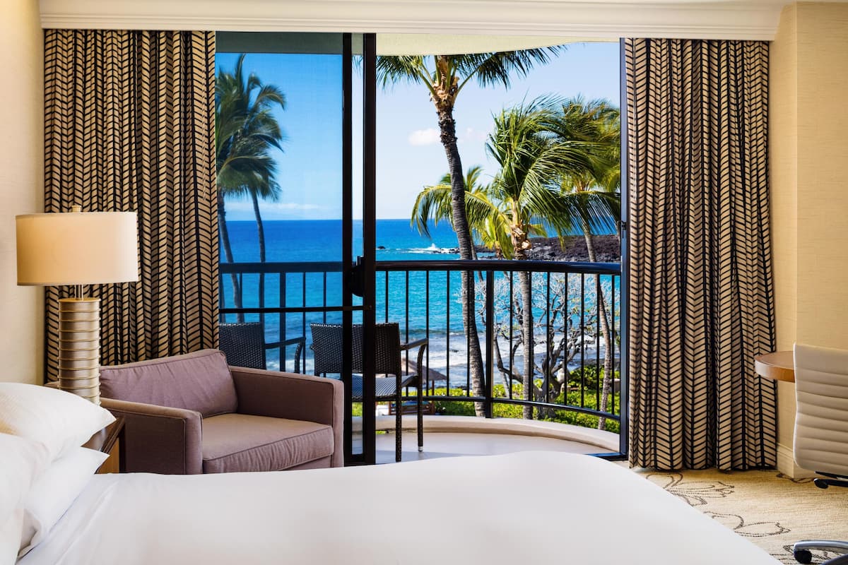 A hotel room with a bed, a chair, a lamp, and a balcony with a view of the ocean and palm trees.
