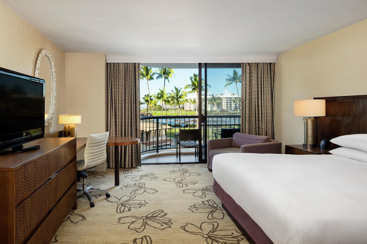Ein Hotelzimmer mit Bett, Schreibtisch mit Fernseher, Stuhl, kleinem Tisch und einem Balkon mit Blick auf Palmen und einen Pool.