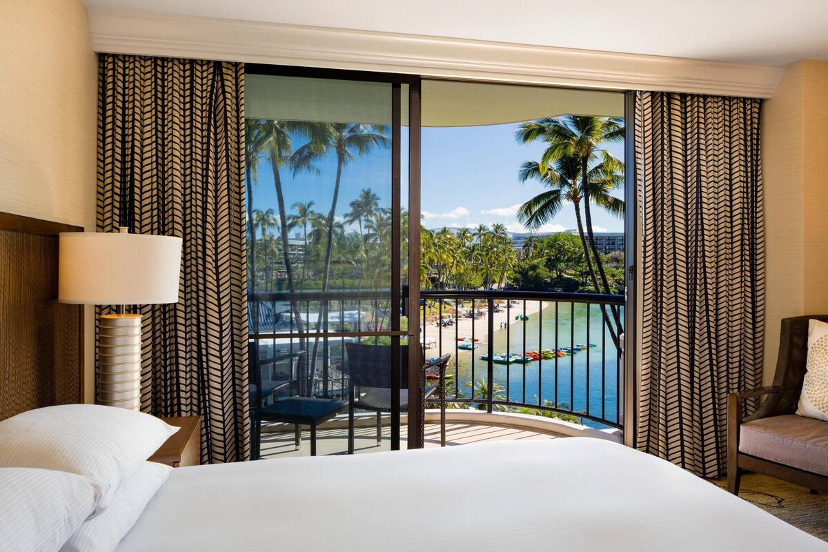 A hotel room with a bed, a chair, a table, and a view of a beach and palm trees through a large window.
