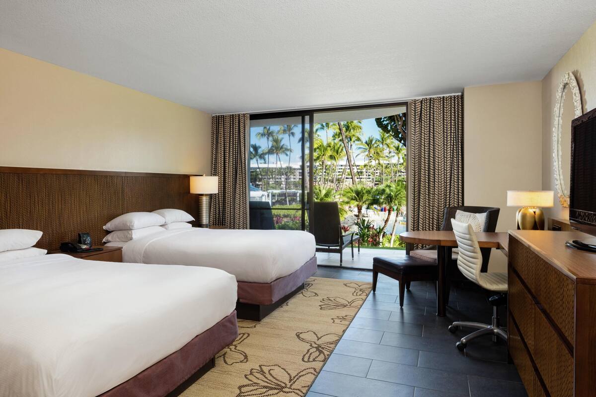 Une chambre d’hôtel avec deux lits, un bureau avec une chaise, une télévision et une vue sur les palmiers et les immeubles.