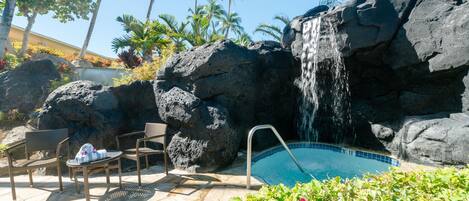 Zona de piscina con cascada, rodeada de rocas y palmeras.