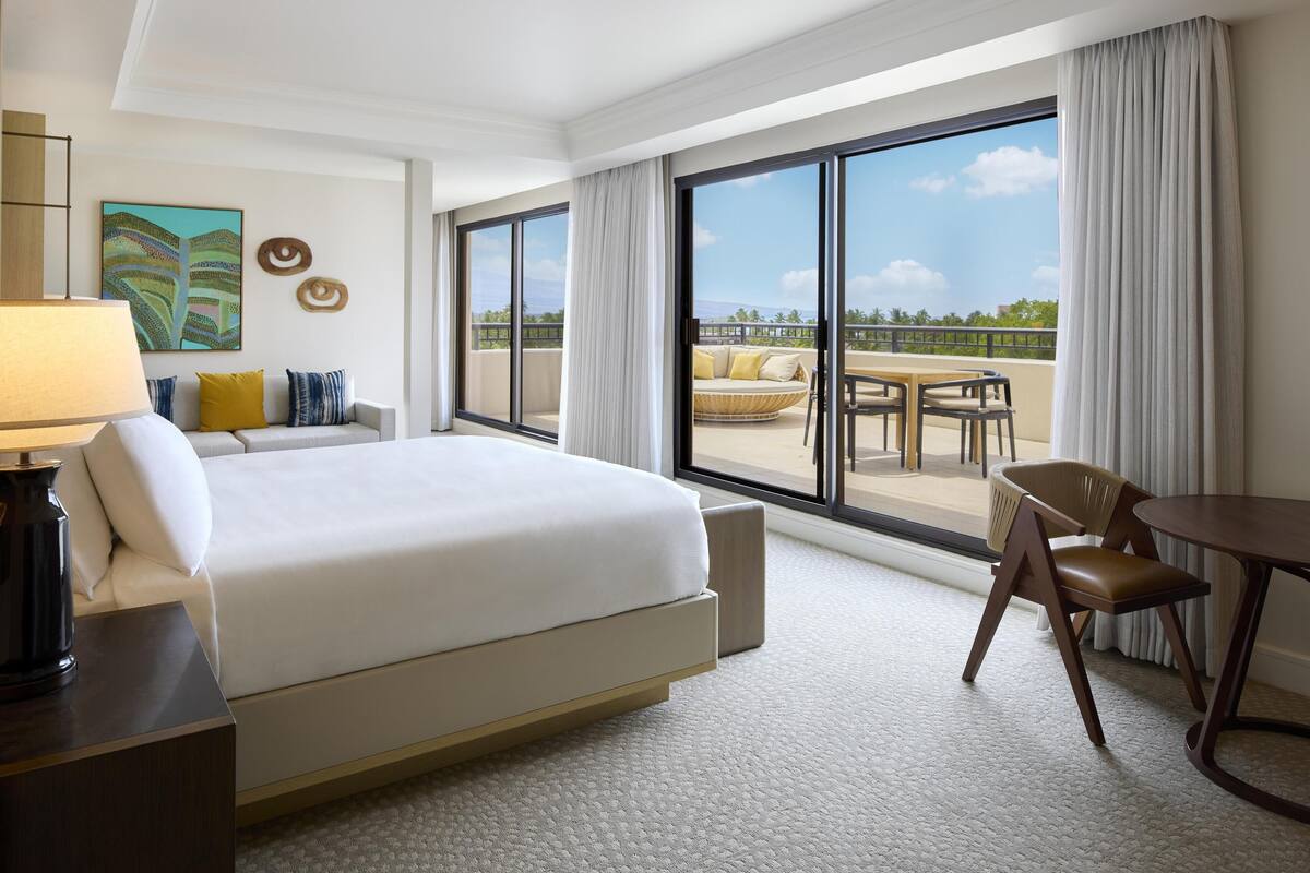A hotel room with a bed, a desk, a chair, a balcony with a table and chairs, and a large window.