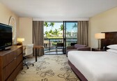 A hotel room with a bed, a desk with a television, a chair, a small table, and a balcony with a view of palm trees and a pool.