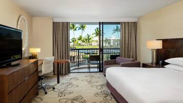 A hotel room with a bed, a desk with a television, a chair, a small table, and a balcony with a view of palm trees and a pool.