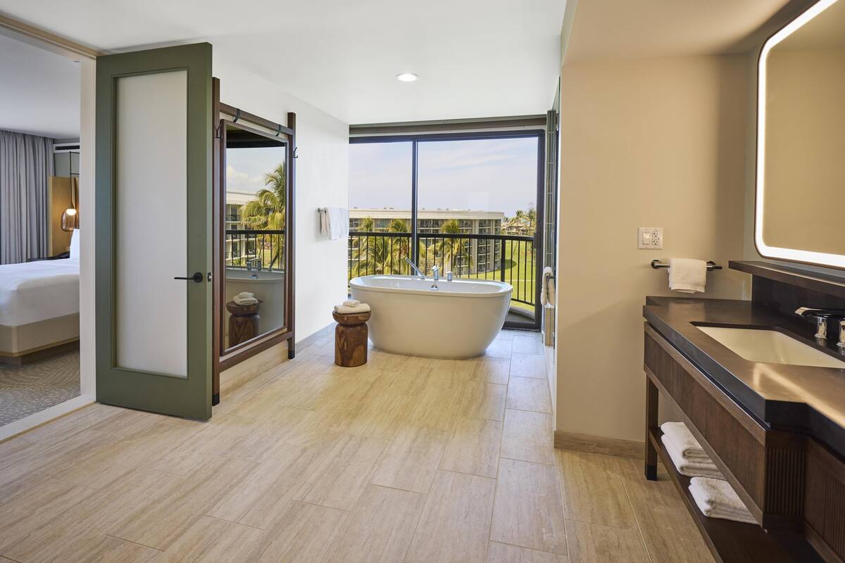 A modern hotel bathroom with a large bathtub, a separate shower area, a vanity with a mirror, and a balcony with a view of palm trees.
