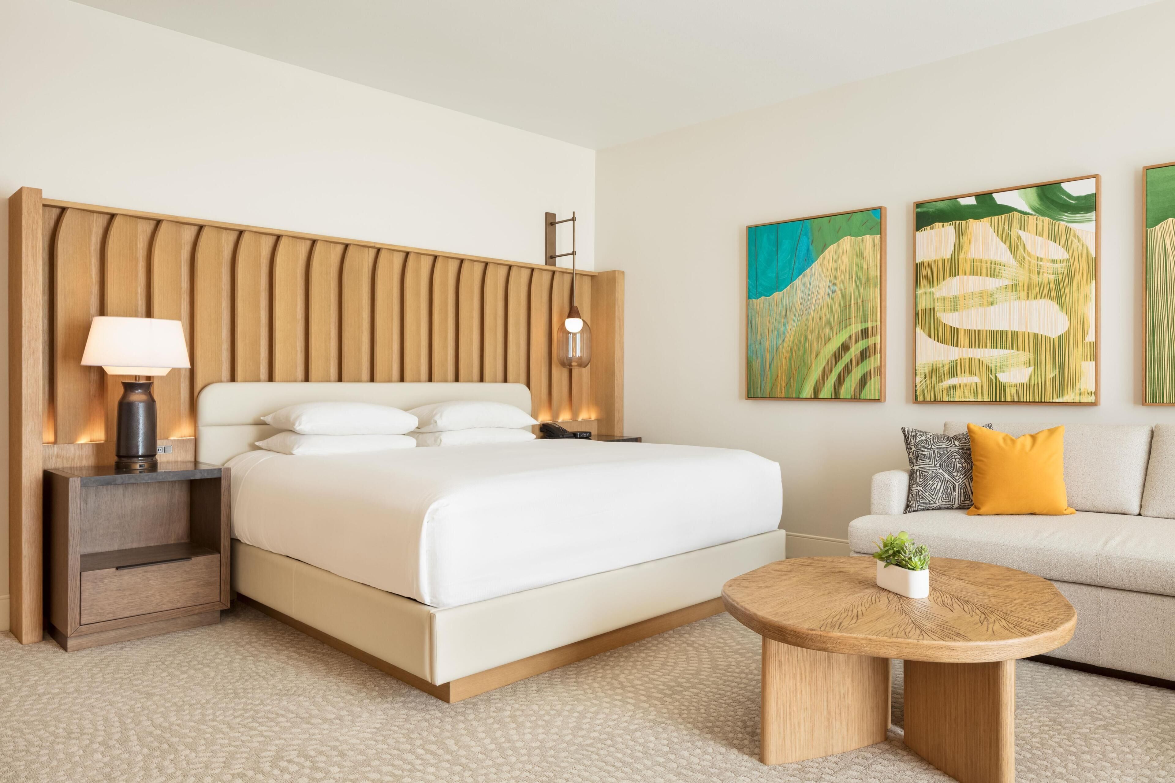 A modern hotel room with a large bed, bedside tables, a round coffee table, and artwork on the walls.