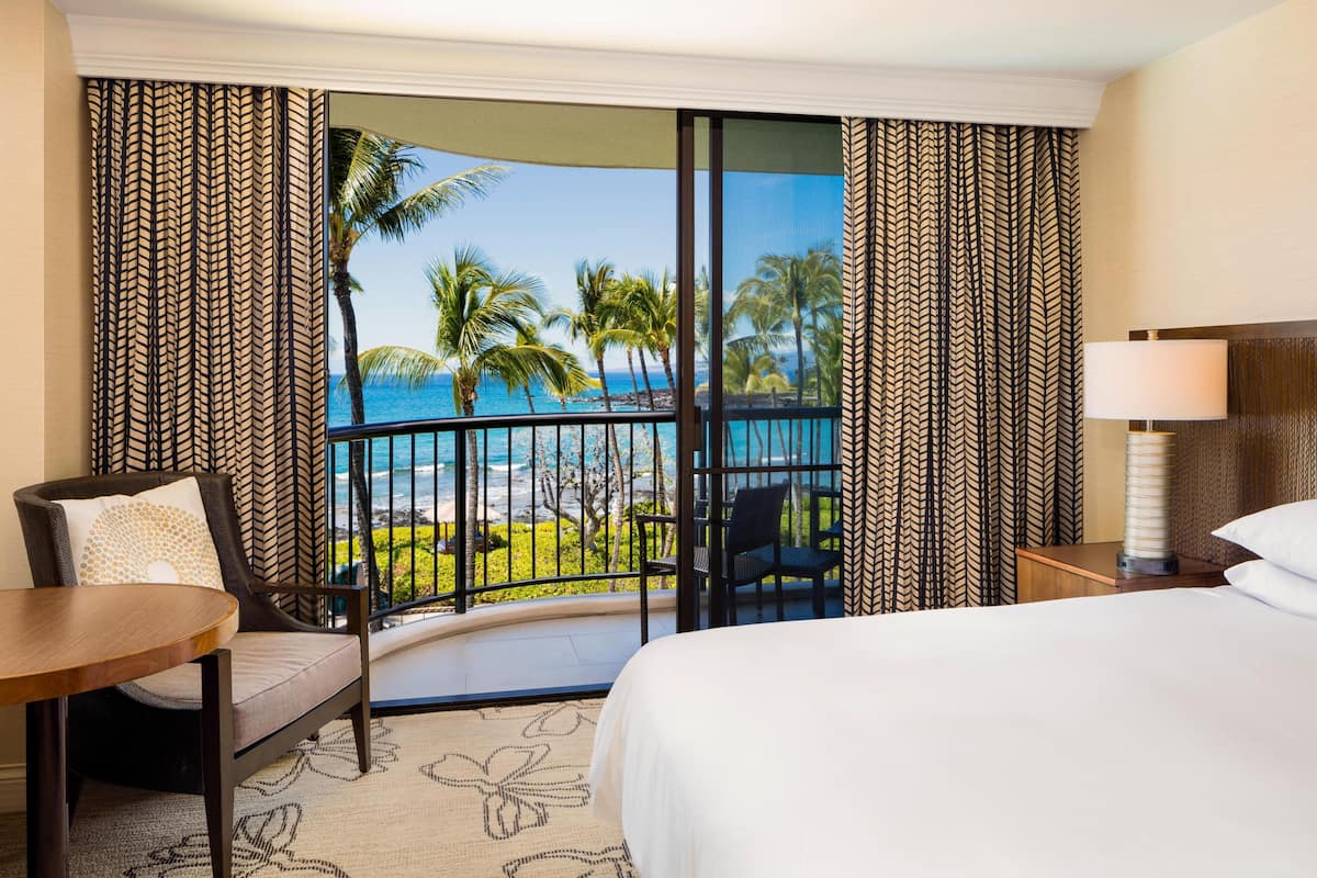 A hotel room with a bed, a chair, a table, a nightstand, and a view of the ocean and palm trees.