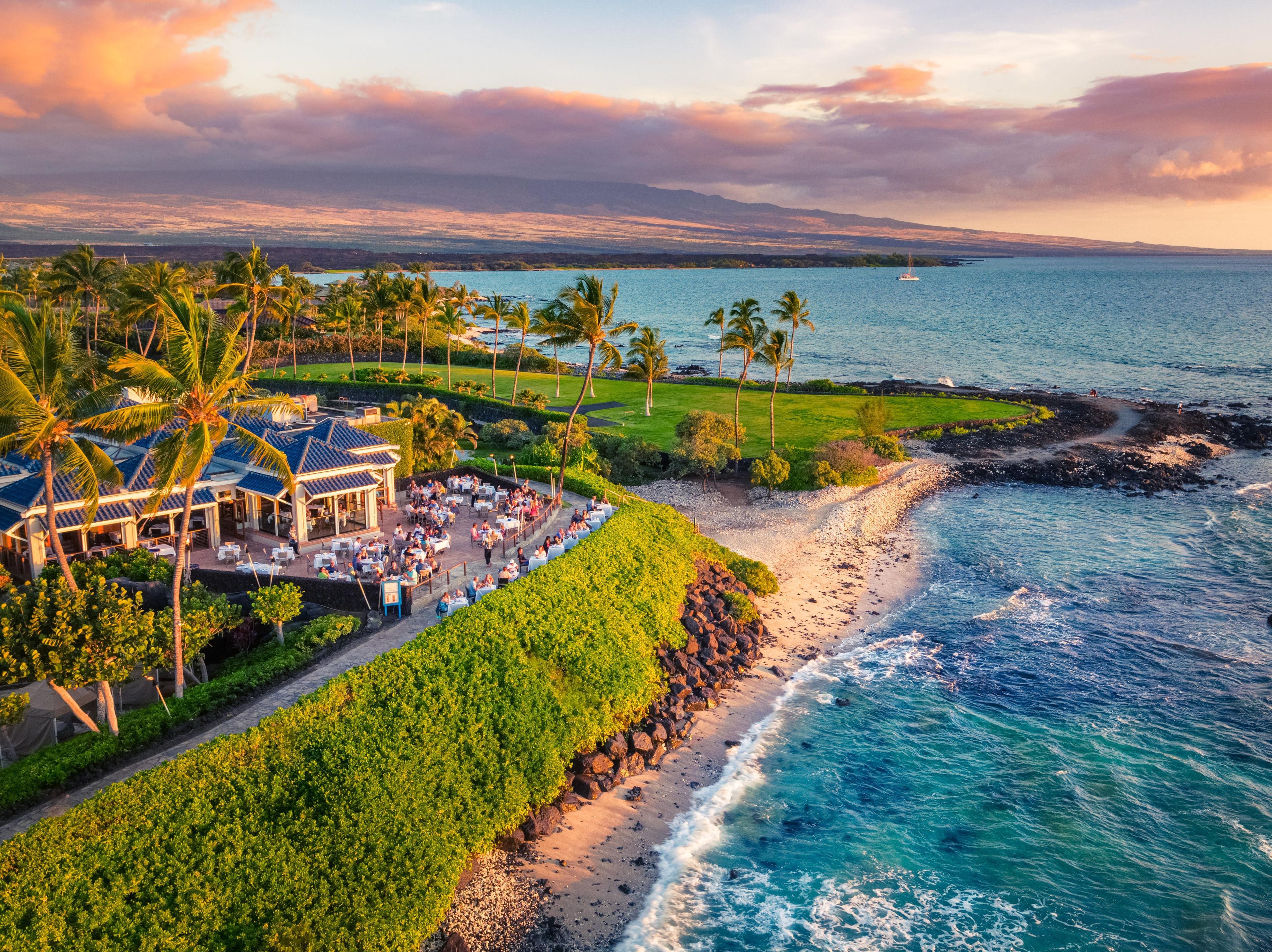 A coastal resort with a dining area, palm trees, and a view of the ocean.