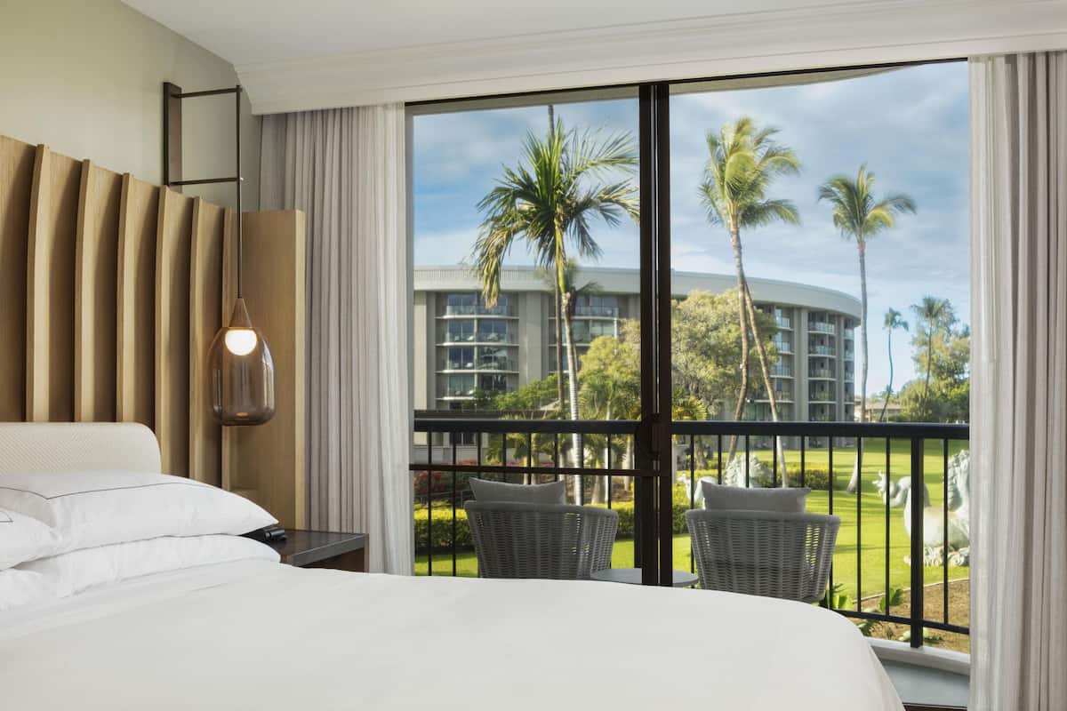 Une chambre d’hôtel avec un grand lit, une vue sur des palmiers et un immeuble, et un balcon équipé de chaises en osier.