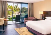 A hotel room with a bed, a desk, a chair, and a view of a beach and palm trees.
