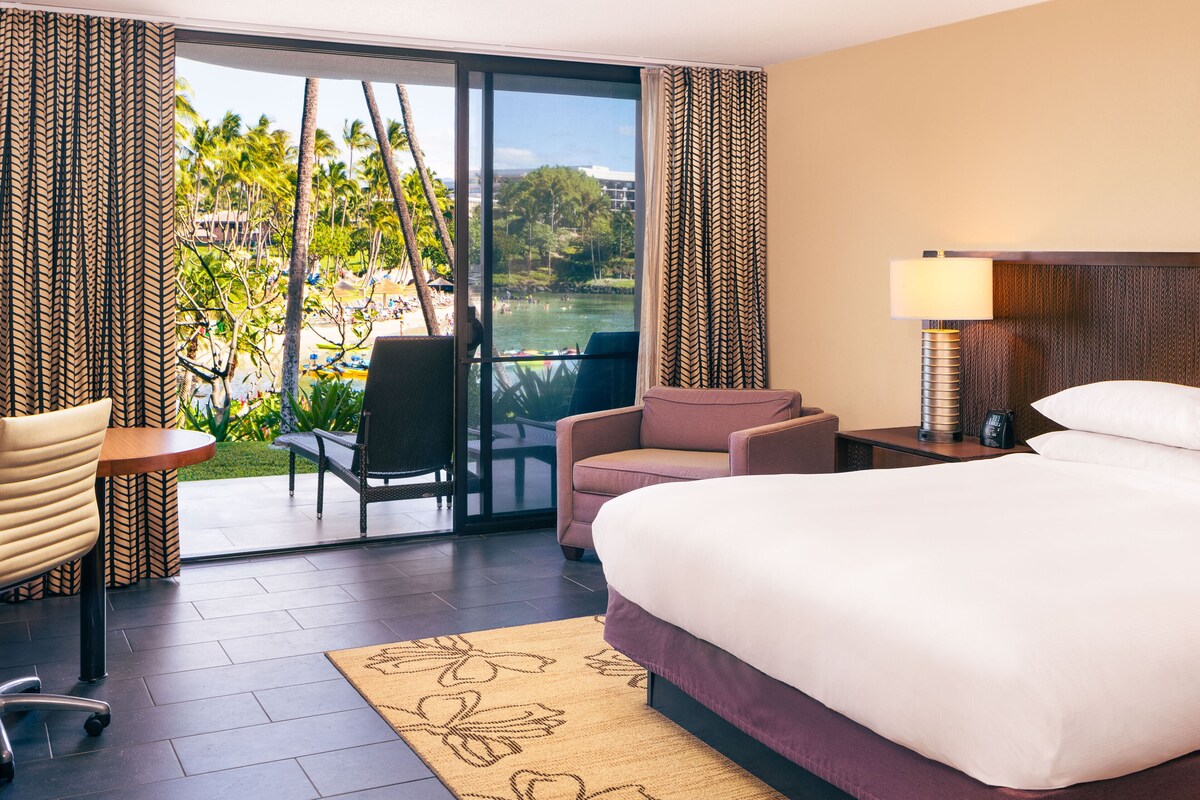 A hotel room with a bed, a desk, a chair, and a view of a beach and palm trees.