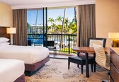 A hotel room with a bed, a desk, a chair, and a view of a pool and palm trees.