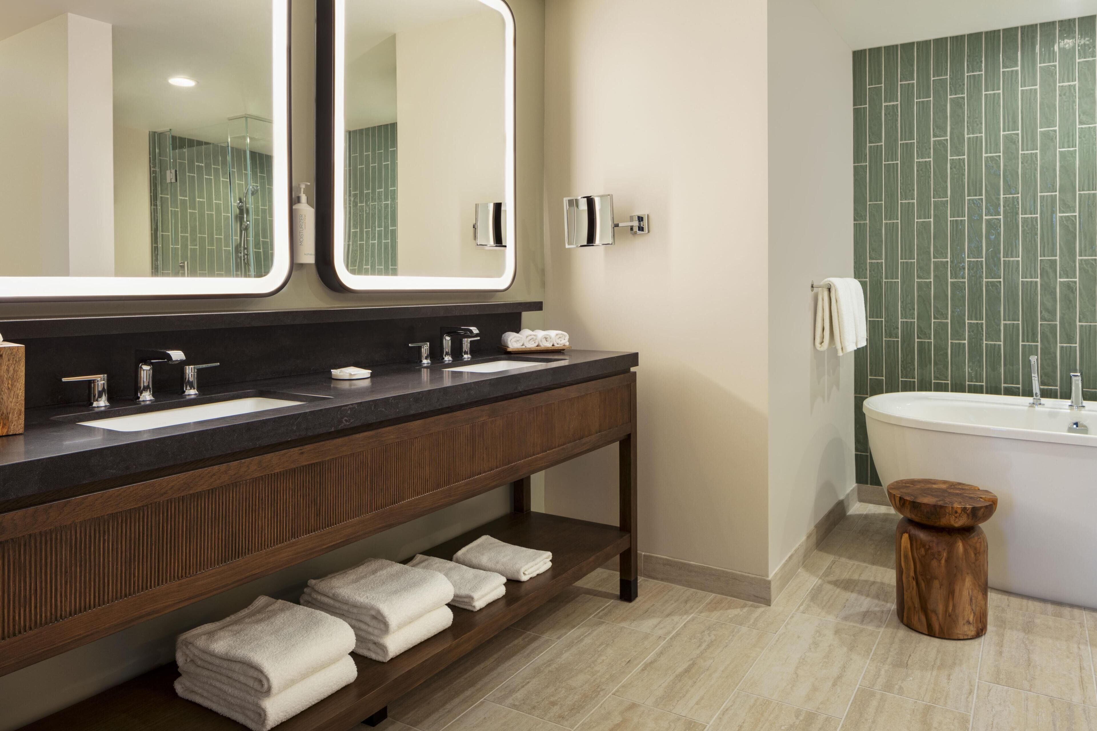 A bathroom with two sinks, a large mirror, a vanity, a bathtub, and a wooden stool.