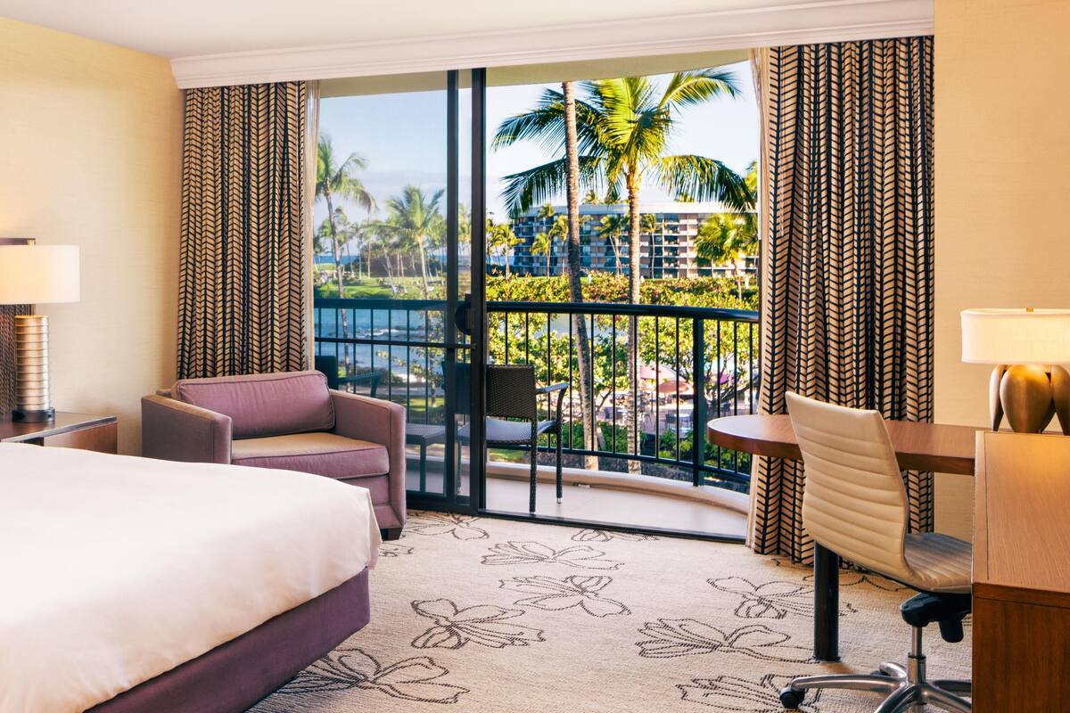 A hotel room with a bed, a desk, a chair, and a view of palm trees and a pool.