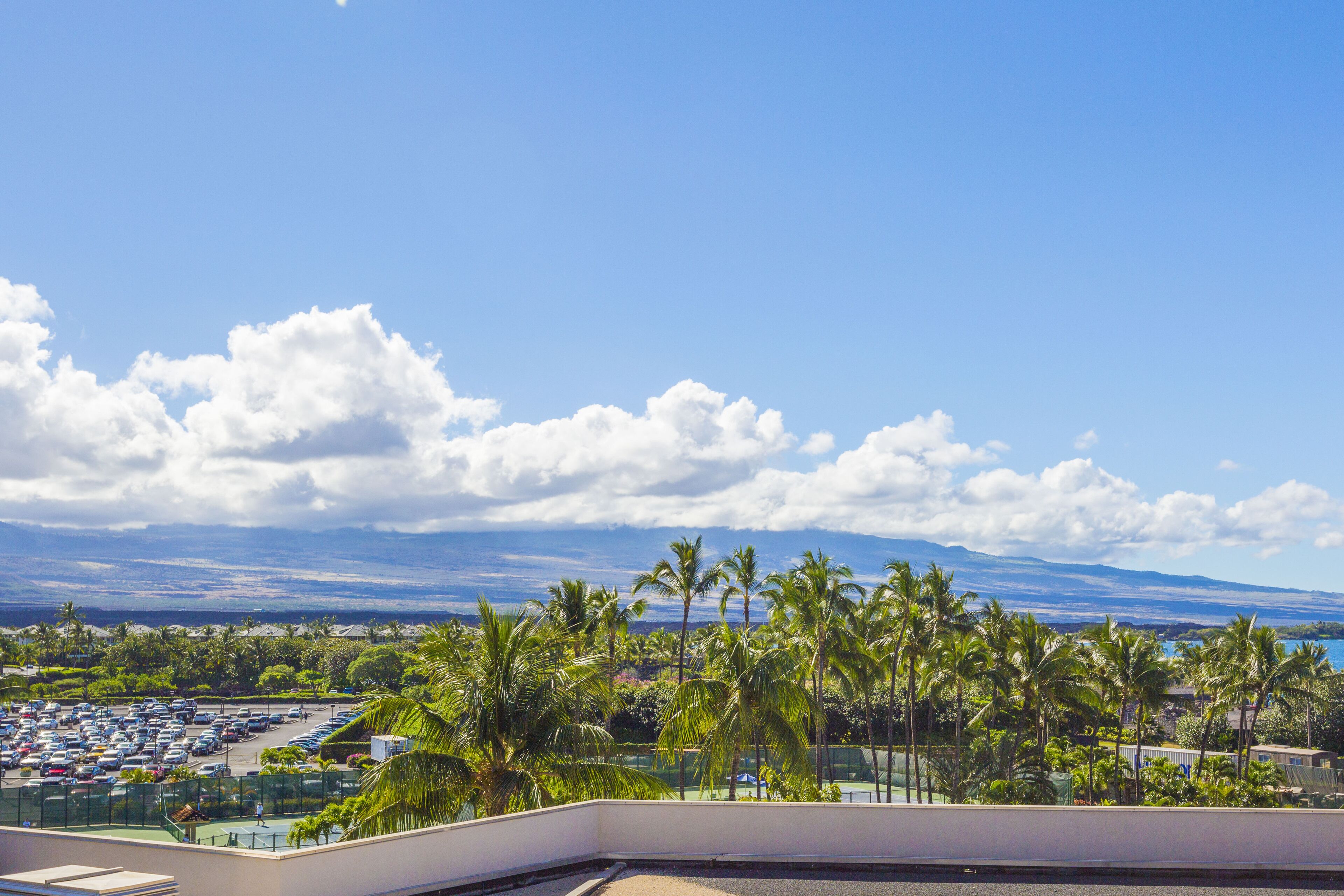 A hotel balcony with a view of a parking lot, palm trees, and a distant mountain range.