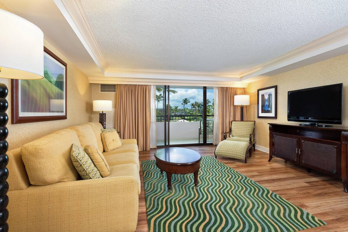 A hotel room with a sofa, armchair, television, and a view of palm trees through the balcony doors.