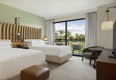 A hotel room with two beds, a desk, a chair, and a balcony with a view of palm trees.