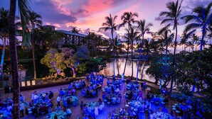 An outdoor dining area with tables and chairs, palm trees, and a body of water in the background.