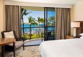 A hotel room with a bed, a chair, a small table, and a view of the ocean with palm trees.