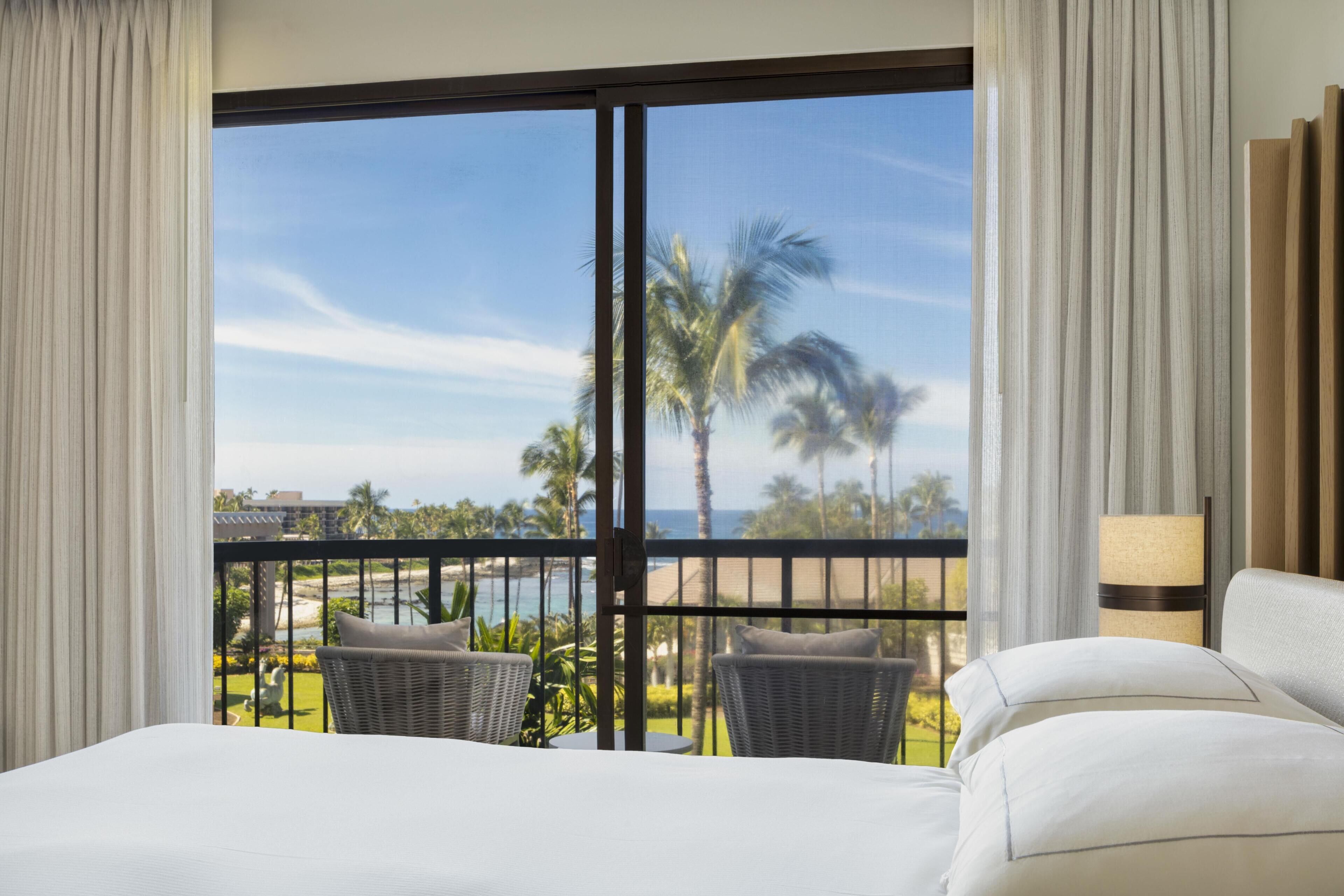 A hotel room with a large window, a bed, two chairs, and a view of the beach and palm trees.