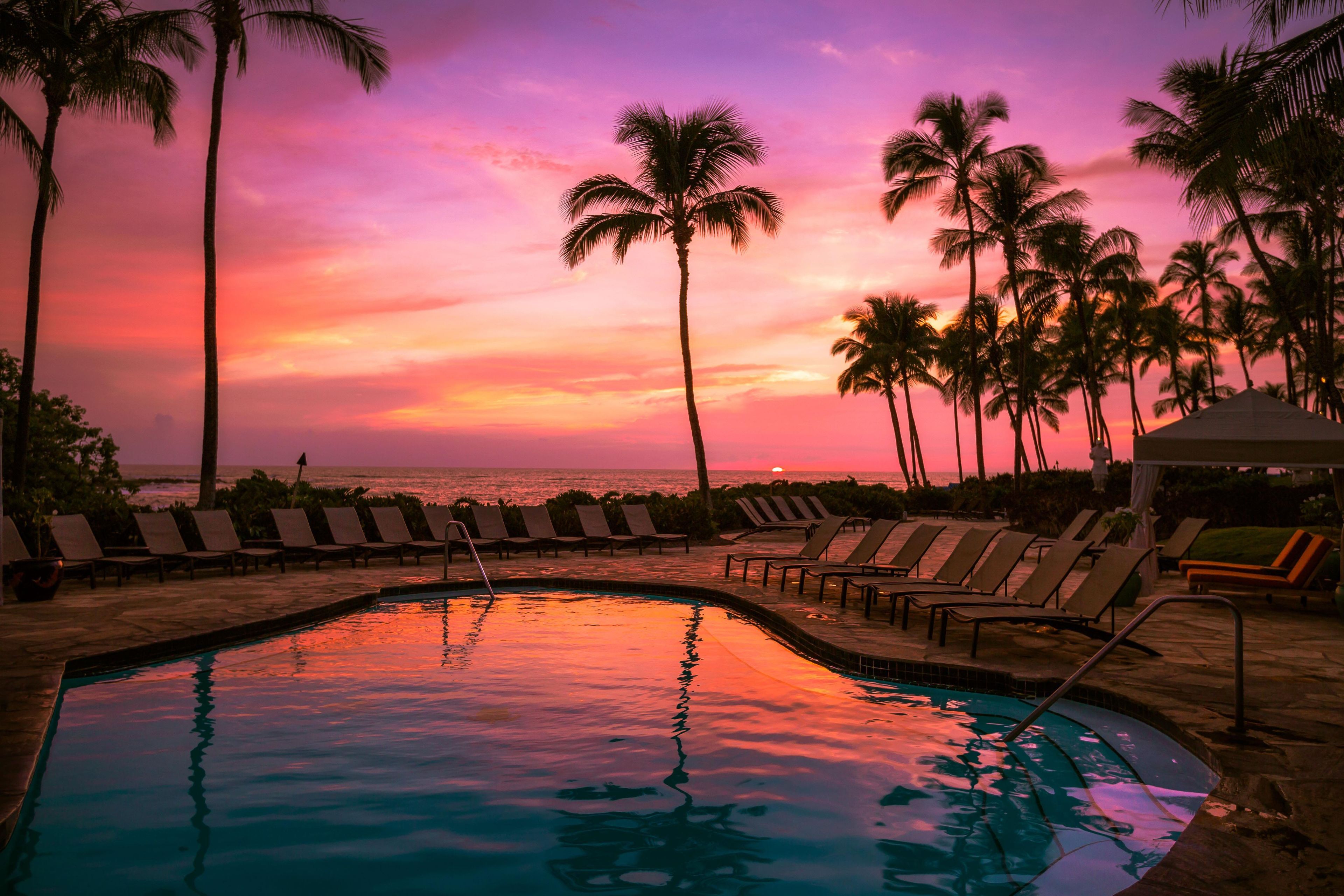 A pool area with lounge chairs and palm trees, overlooking the ocean at sunset.