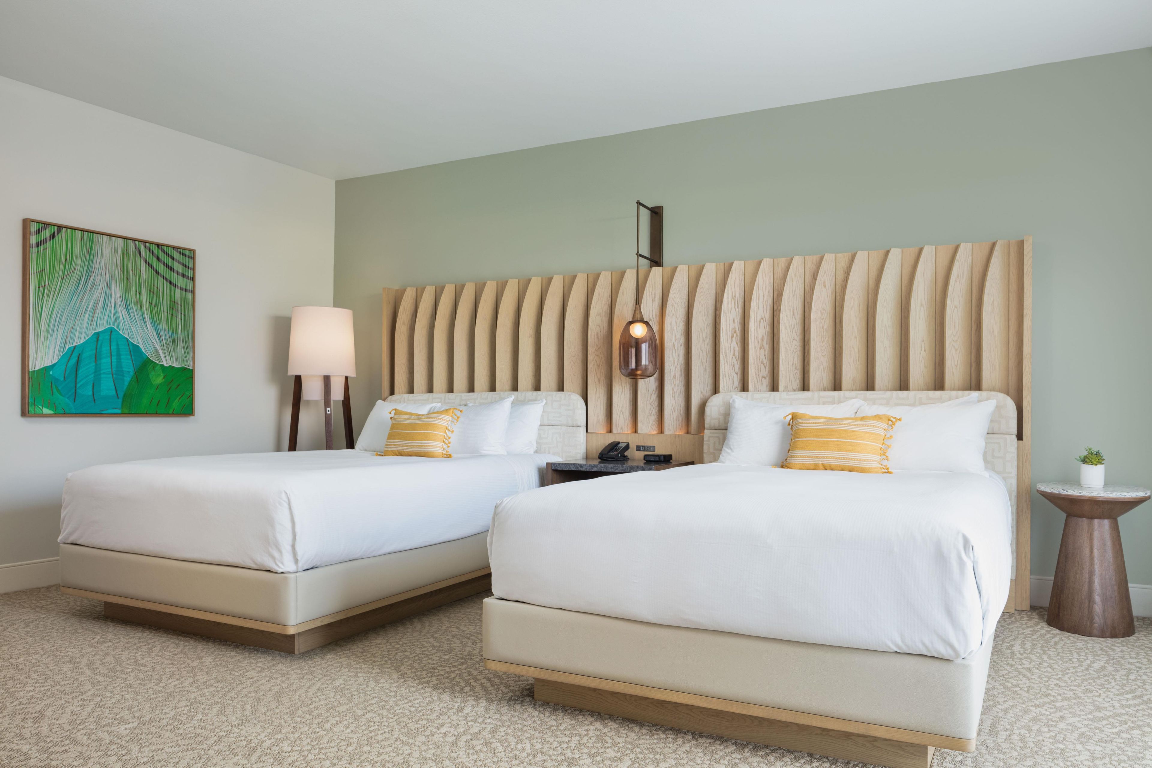 A hotel room with two beds, a painting on the wall, a bedside lamp, a small table with a plant, and a wall-mounted light fixture.