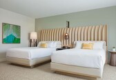 A hotel room with two beds, a painting on the wall, a bedside lamp, a small table with a plant, and a wall-mounted light fixture.