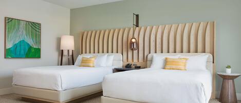 A hotel room with two beds, a painting on the wall, a bedside lamp, a small table with a plant, and a wall-mounted light fixture.