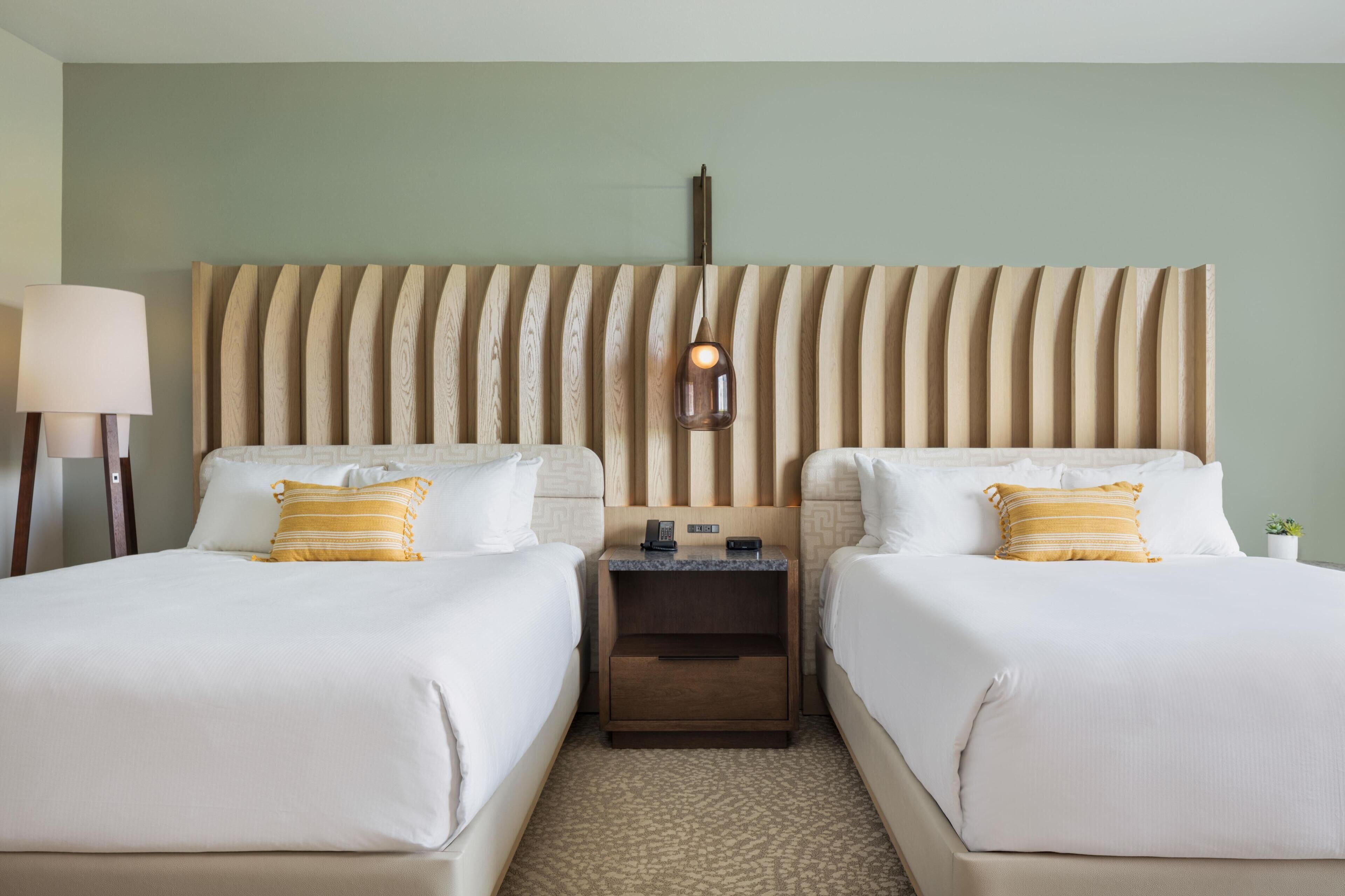 A hotel room with two beds, a nightstand, a lamp, and a wall-mounted light fixture.